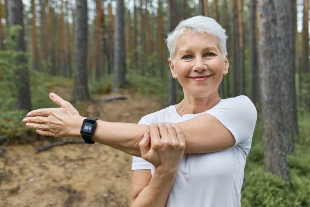 Healthy senior woman stretching her arm outdoors in nature, promoting wellness, fitness, and longevity with Dr. Thind's Homeopathic Clinic.